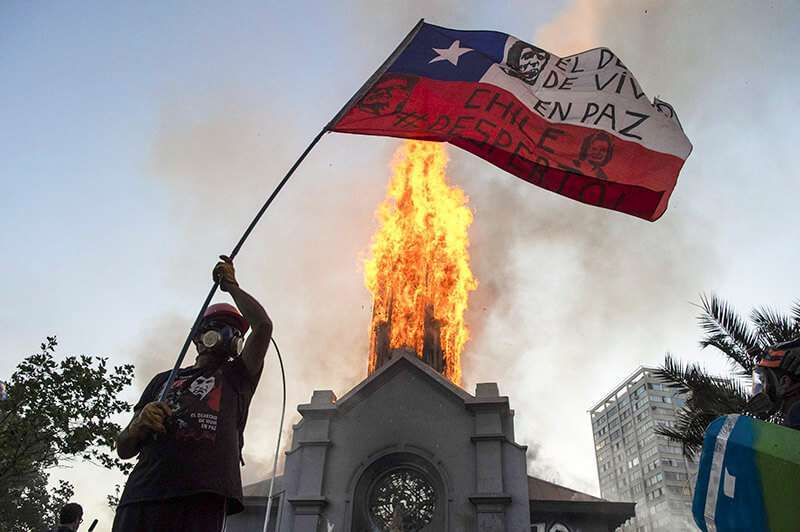 Atalayar_ Un manifestante ondea una bandera chilena frente a la iglesia en llamas de Asunción, en el primer aniversario del levantamiento social