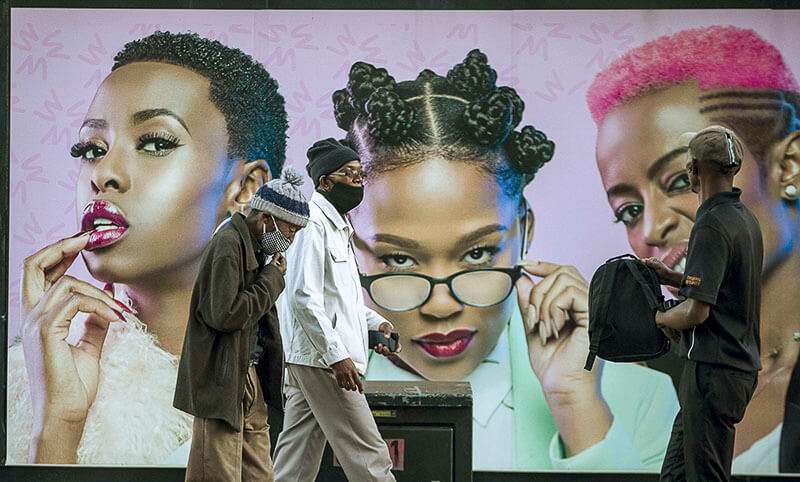 Hombres que usan una máscara facial para protegerse contra el coronavirus, pasan frente a un cartel publicitario en Soweto, Sudáfrica, el lunes 29 de junio de 2020
