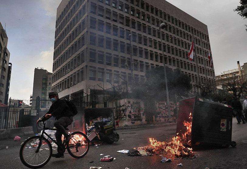 PHOTO/AP  -   Un hombre pasa con su bicicleta por delante de los contenedores de basura incendiados por los manifestantes que bloquean una carretera principal frente al edificio del Banco Central del Líbano